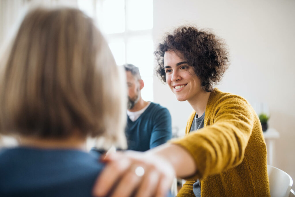 Een vrouw legt glimlachend haar hand op de schouder van een andere vrouw tijdens een groepsgesprek.