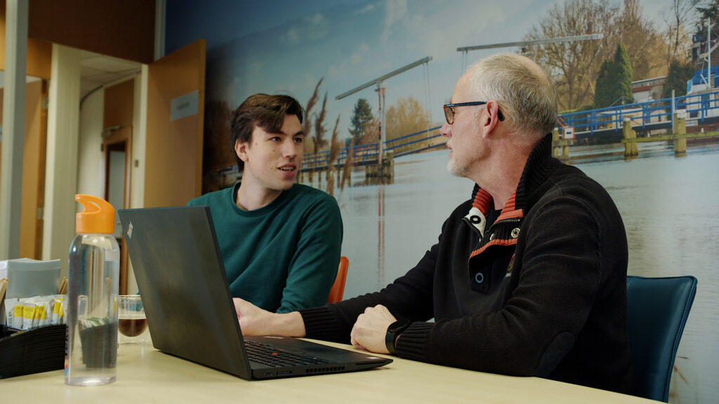 Twee mannen in overleg aan tafel, met een laptop open voor zich.